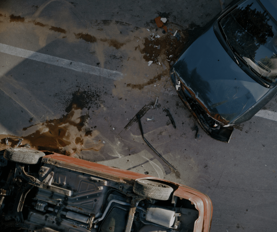 Bird's eye view of two cars in an accident. An orange one is flipped over and the black one has a crushed front. Dirt a and debris sprinkle the ground.